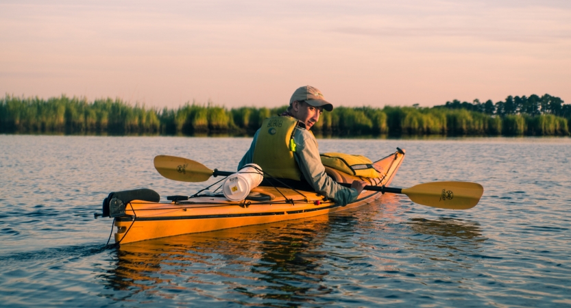 A young person takes a break from paddling a kayak to look back at the camera. There is a line of trees in the background. 